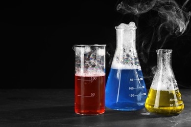 Photo of Laboratory glassware with colorful liquids and steam on dark table against black background, space for text. Chemical reaction