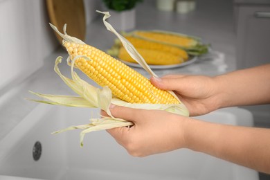 Photo of Woman husking corn cob in kitchen, closeup