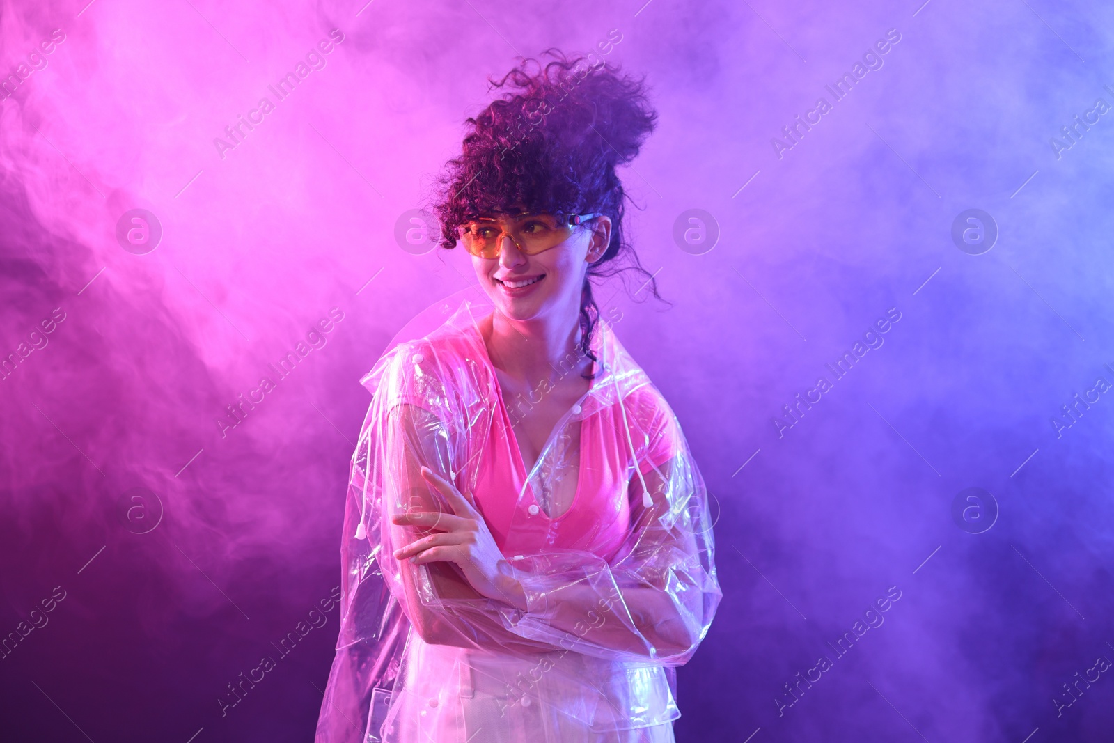 Photo of Beautiful young woman in transparent coat and sunglasses posing on color background in neon lights and smoke