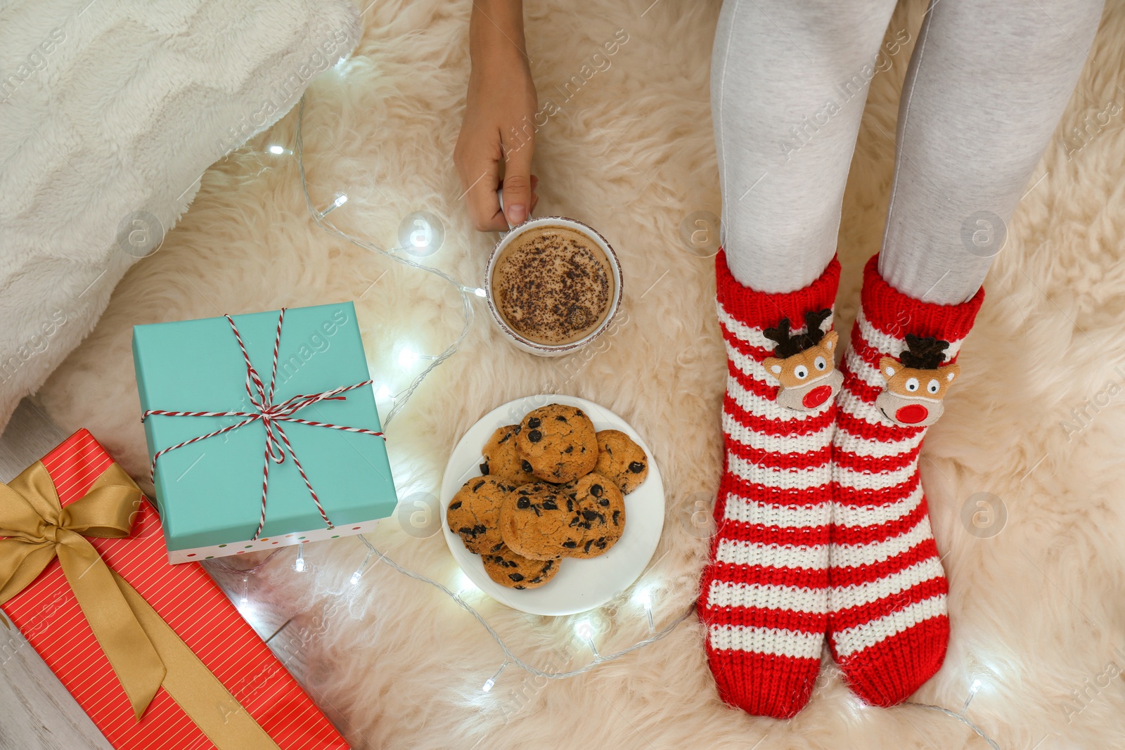 Photo of Woman with cup of hot drink, cookies and gifts on white fuzzy carpet, top view. Christmas season