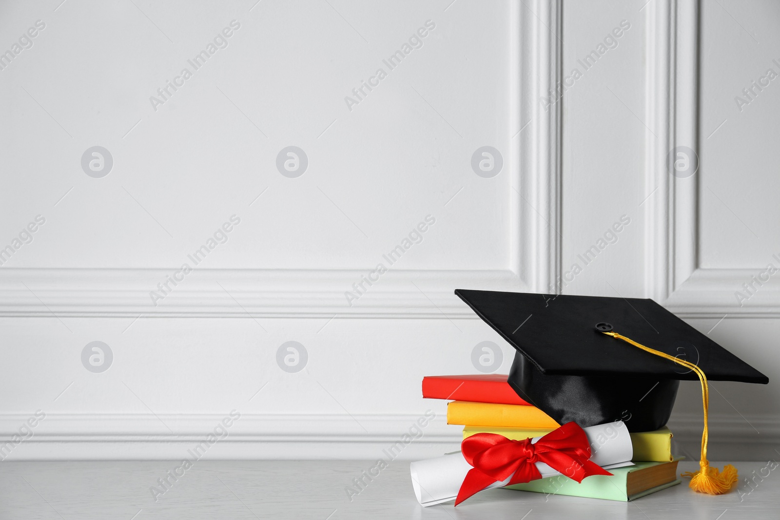 Photo of Graduation hat, books and diploma on floor near white wall, space for text