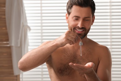 Photo of Smiling man dropping cosmetic serum onto his hand in bathroom. Space for text