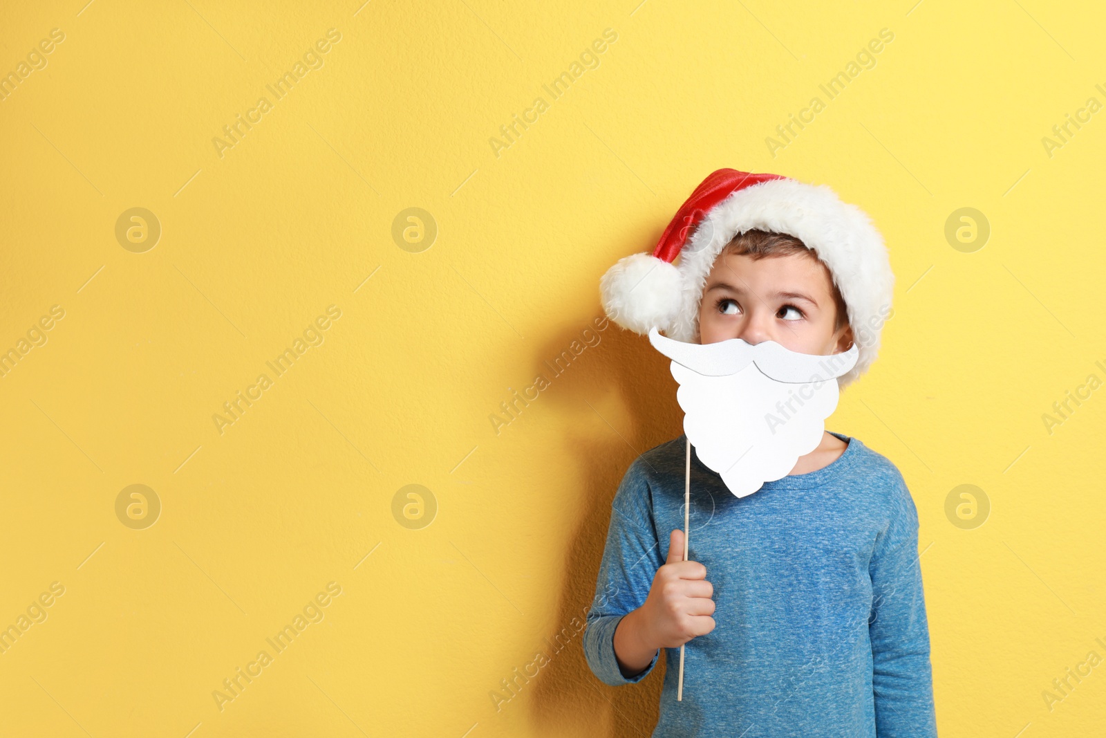 Image of Cute little boy with Santa hat and white beard prop on yellow background, space for text. Christmas celebration