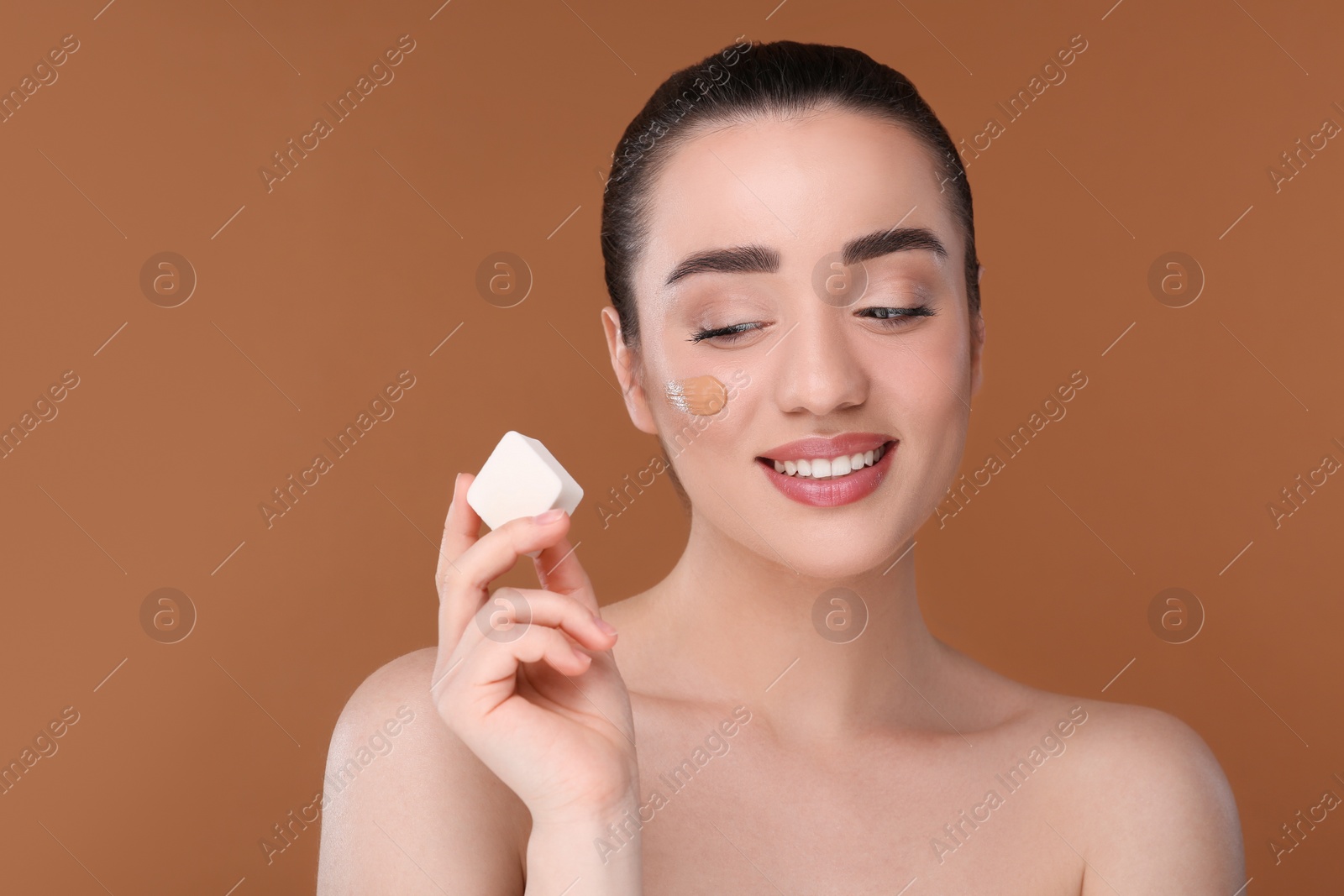 Photo of Woman applying foundation on face with makeup sponge against brown background