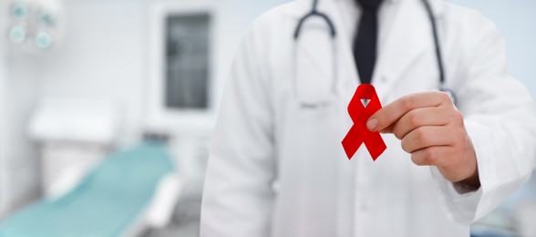 Cancer awareness. Doctor holding red ribbon on blurred background, closeup. Banner design with space for text