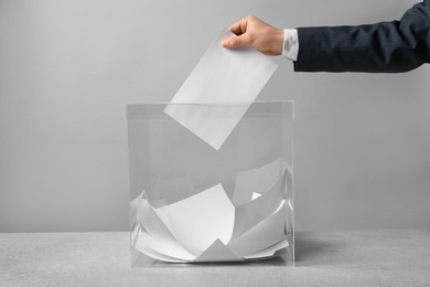 Photo of Man putting his vote into ballot box on table against light background, closeup