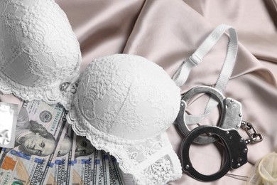 Photo of Prostitution concept. Handcuffs, dollar banknotes, condoms and bra on beige silk cloth, flat lay
