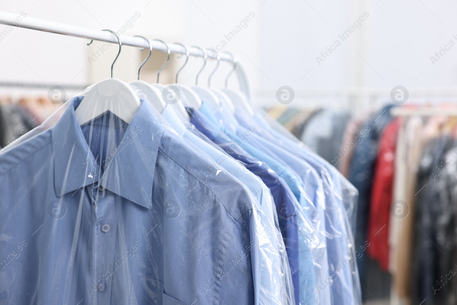 Photo of Dry-cleaning service. Many different clothes in plastic bags hanging on rack indoors, closeup