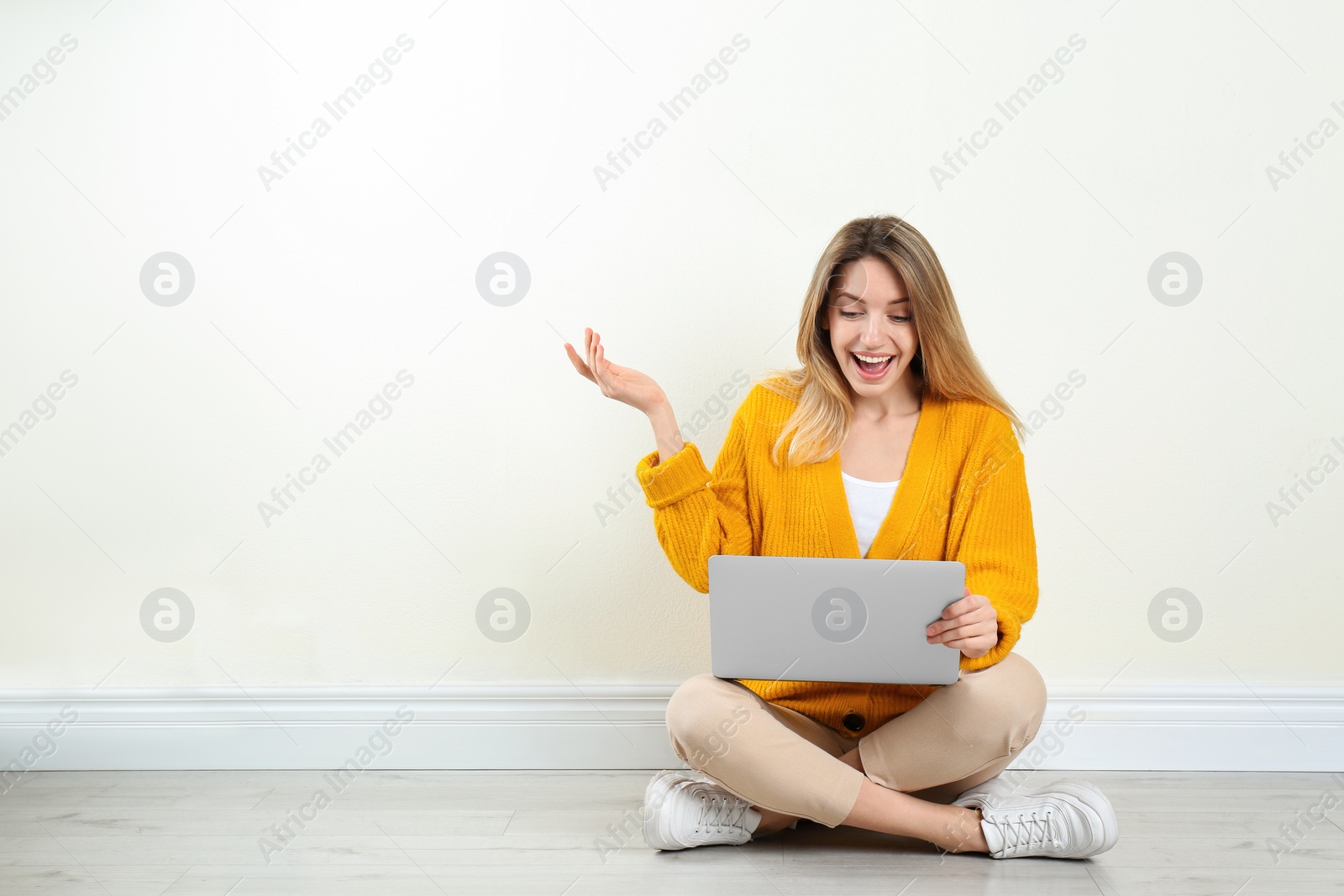 Photo of Happy young woman with laptop sitting on floor near light wall indoors. Space for text