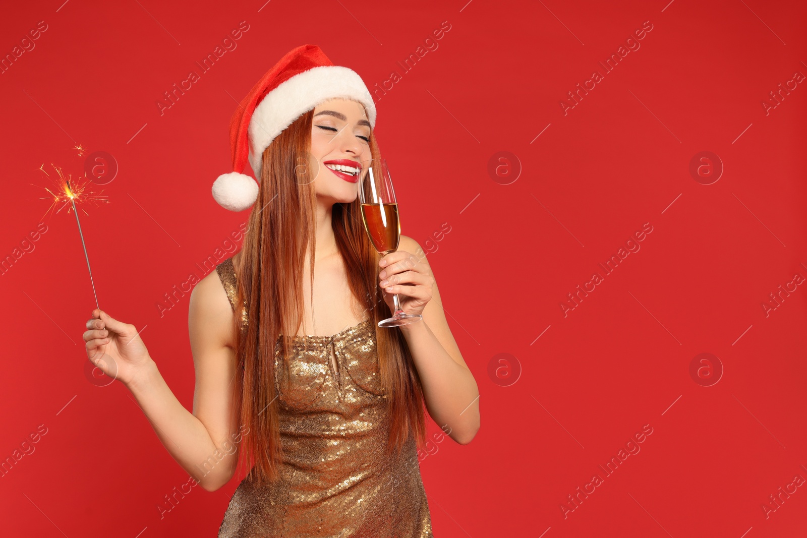 Photo of Young woman in Santa hat with burning sparkler and glass of wine on red background, space for text. Christmas celebration