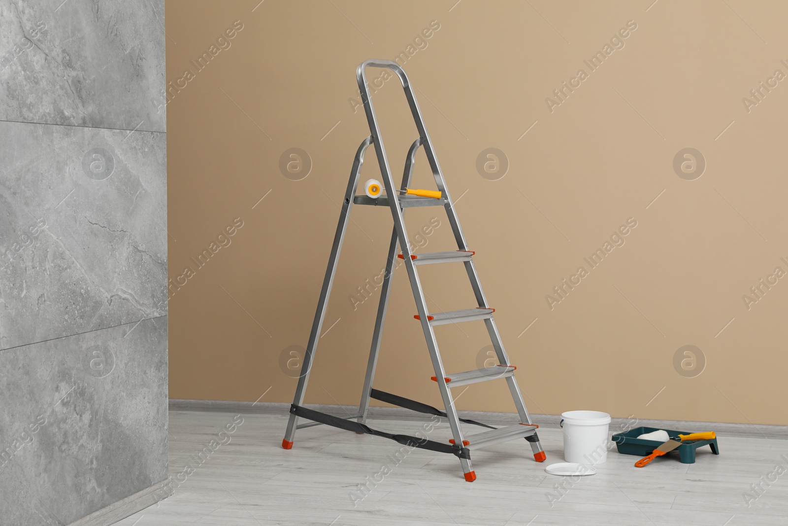 Photo of Metallic folding ladder and painting tools near beige wall indoors