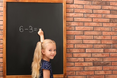 Photo of Cute little left-handed girl doing sums on chalkboard near brick wall