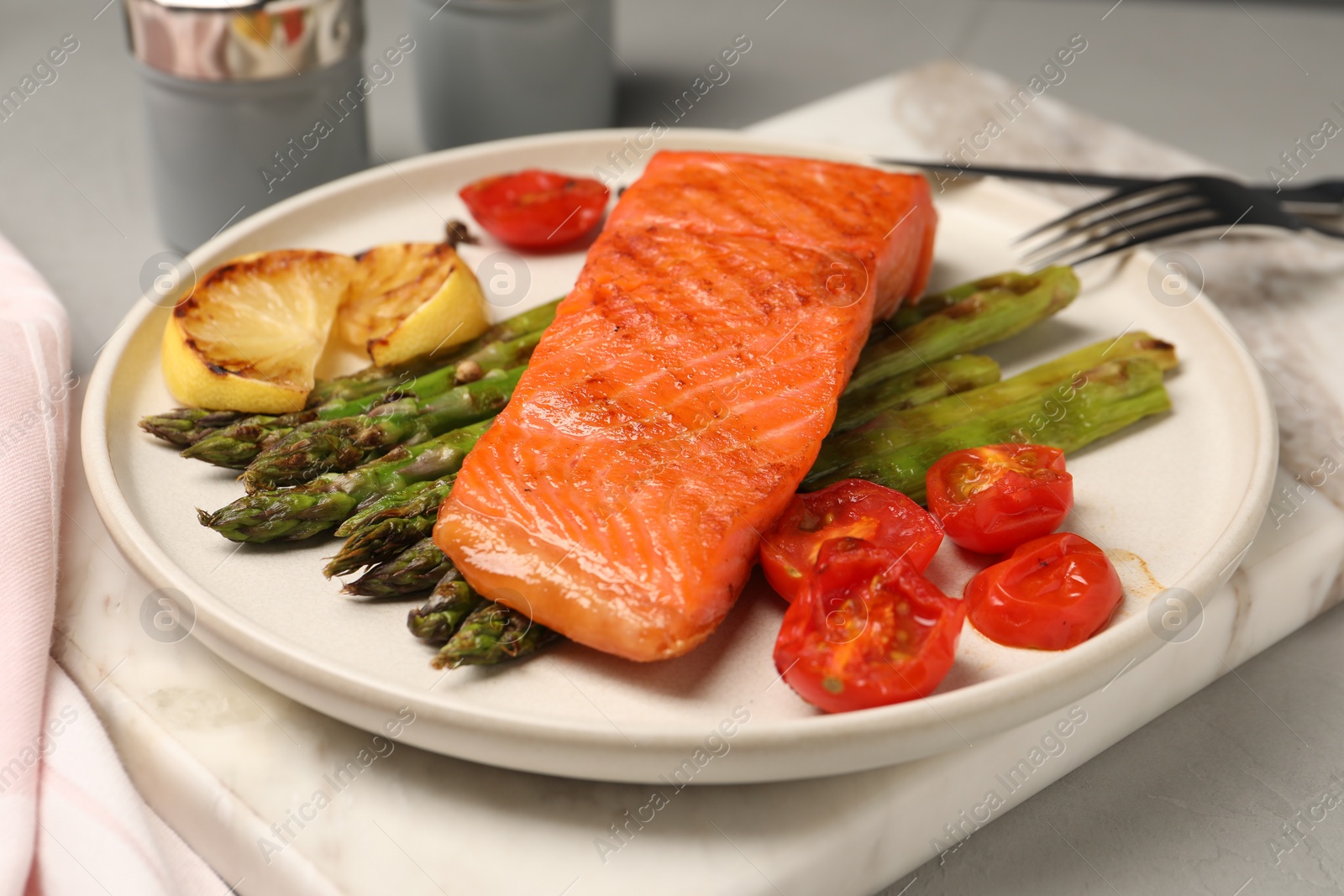 Photo of Tasty grilled salmon with asparagus, tomatoes and lemon on light grey table, closeup