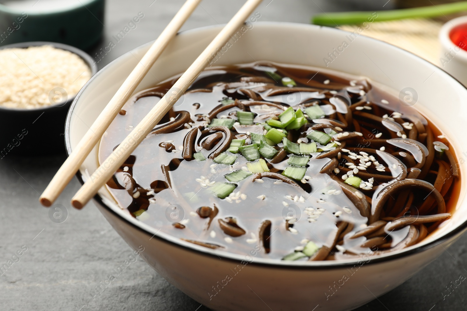 Photo of Tasty soup with buckwheat noodles (soba) and onion in bowl served on grey table, closeup