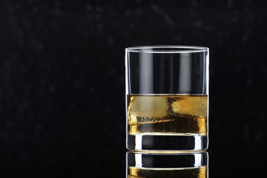 Photo of Tasty whiskey and ice cubes in glass on mirror table against black background, closeup. Space for text