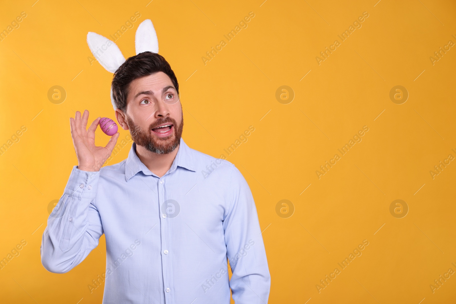 Photo of Emotional man in cute bunny ears headband holding Easter egg on orange background. Space for text