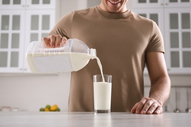 Man pouring milk from gallon bottle into glass at white marble table in kitchen, closeup