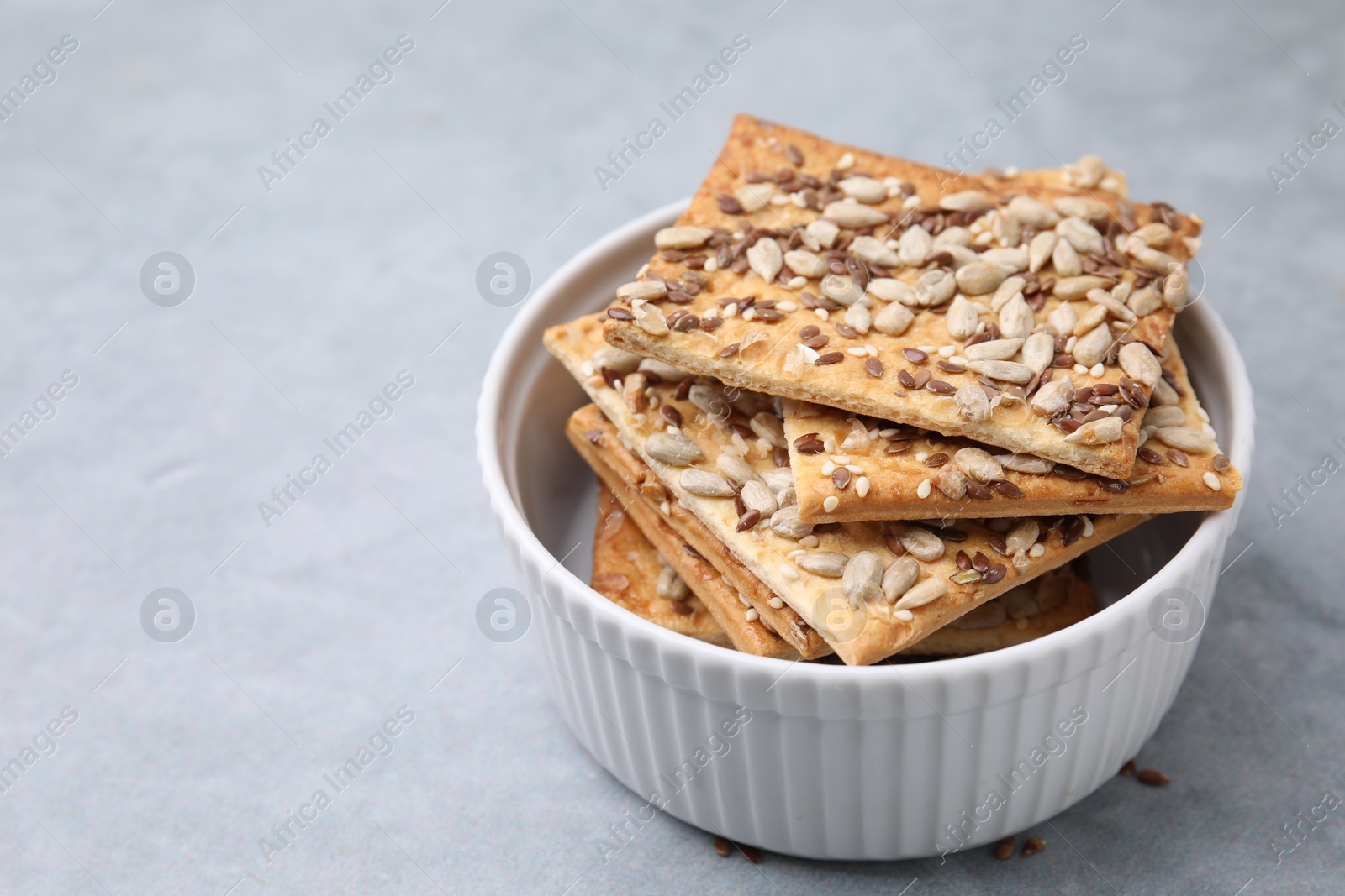 Photo of Cereal crackers with flax, sunflower and sesame seeds in bowl on grey table, closeup. Space for text