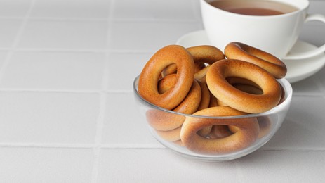 Photo of Bowl with tasty dry bagels (sushki) and cup of tea on white tiled table, space for text
