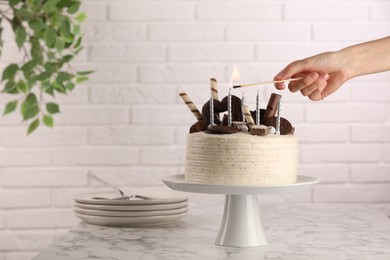 Woman lighting candle on delicious cake decorated with sweets at white marble table, closeup. Space for text