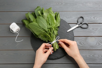 Photo of Woman tying bunch of sorrel leaves at grey wooden table, top view
