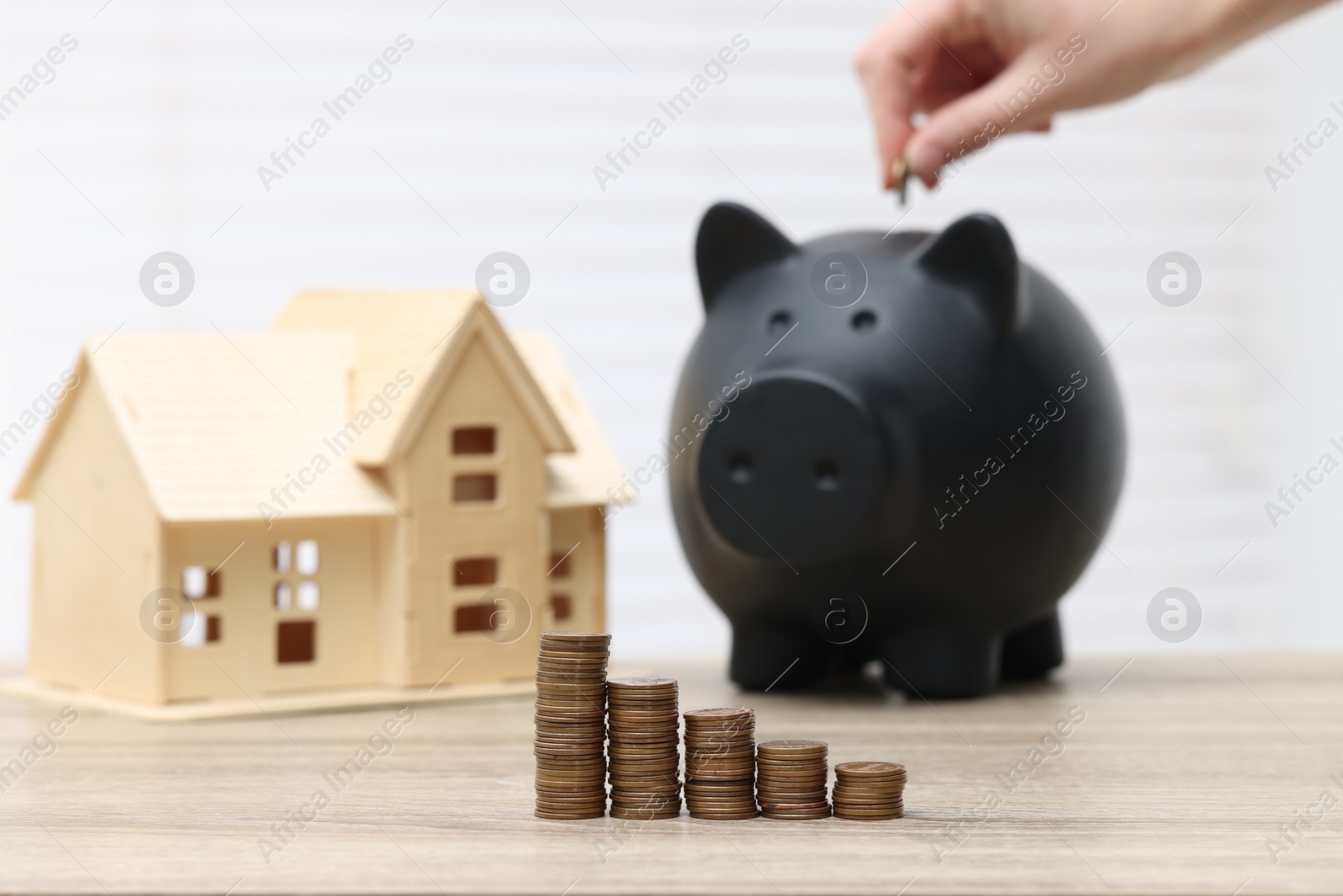 Photo of Woman putting coin into piggy bank at wooden table, focus on stacked coins