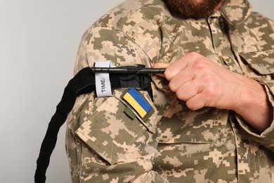 Photo of Ukrainian soldier in military uniform applying medical tourniquet on arm against light grey background, closeup