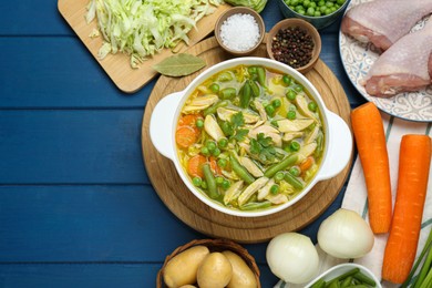 Saucepan of delicious vegetable soup with chicken and different ingredients on blue wooden table, flat lay. Space for text