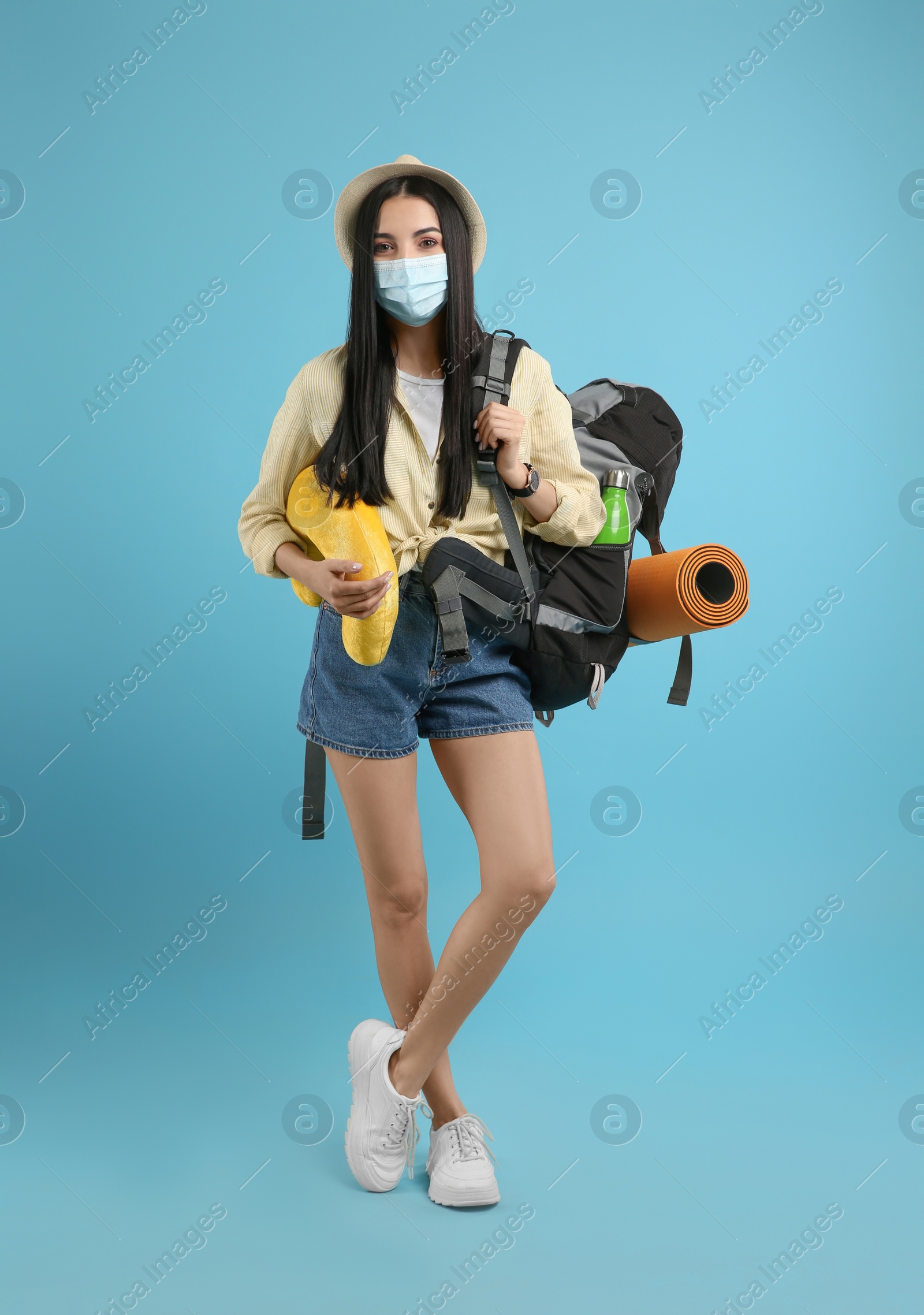 Photo of Female tourist in medical mask with backpack and travel pillow on light blue background