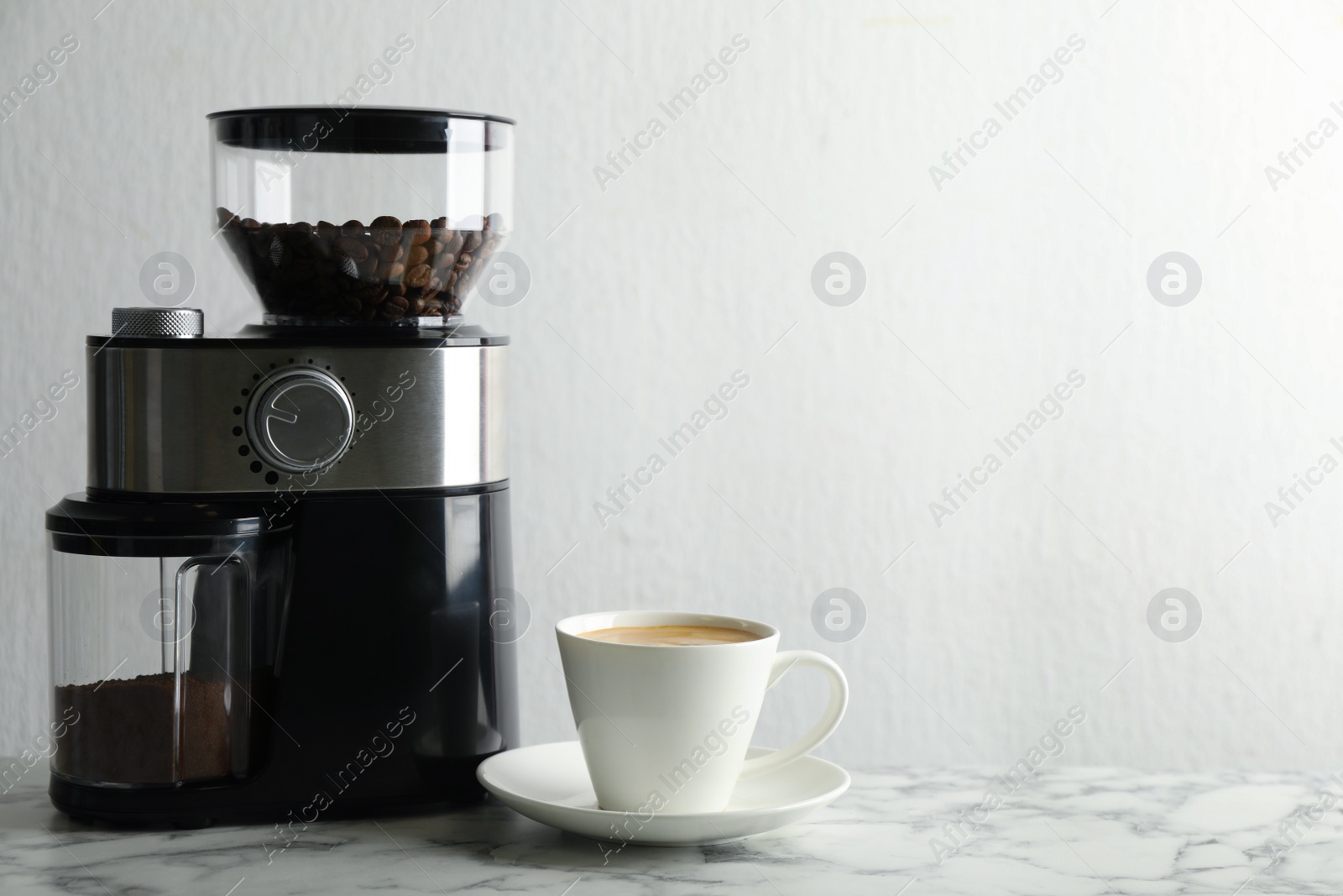 Photo of Modern electric coffee grinder and cup of fresh drink on white marble table. Space for text