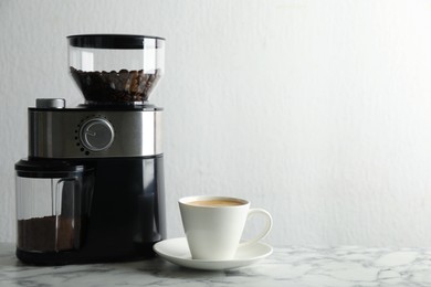 Modern electric coffee grinder and cup of fresh drink on white marble table. Space for text