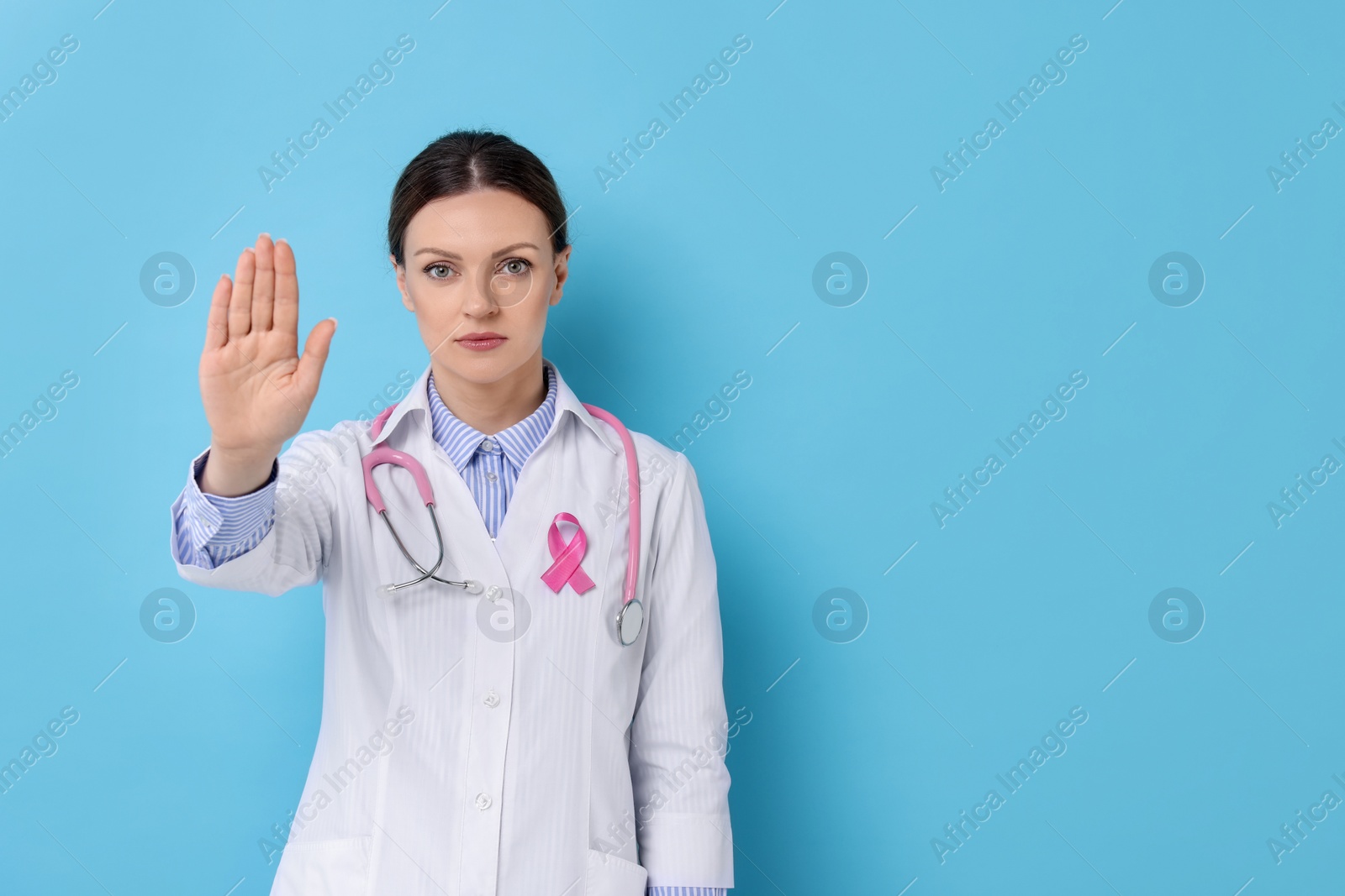 Photo of Mammologist with pink ribbon showing stop gesture against light blue background, space for text. Breast cancer awareness