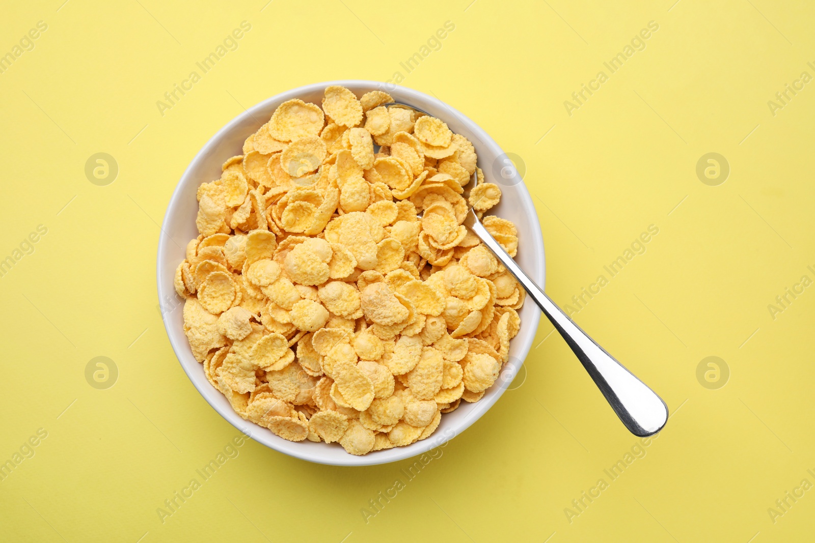 Photo of Breakfast cereal. Tasty corn flakes in bowl and spoon on yellow table, top view