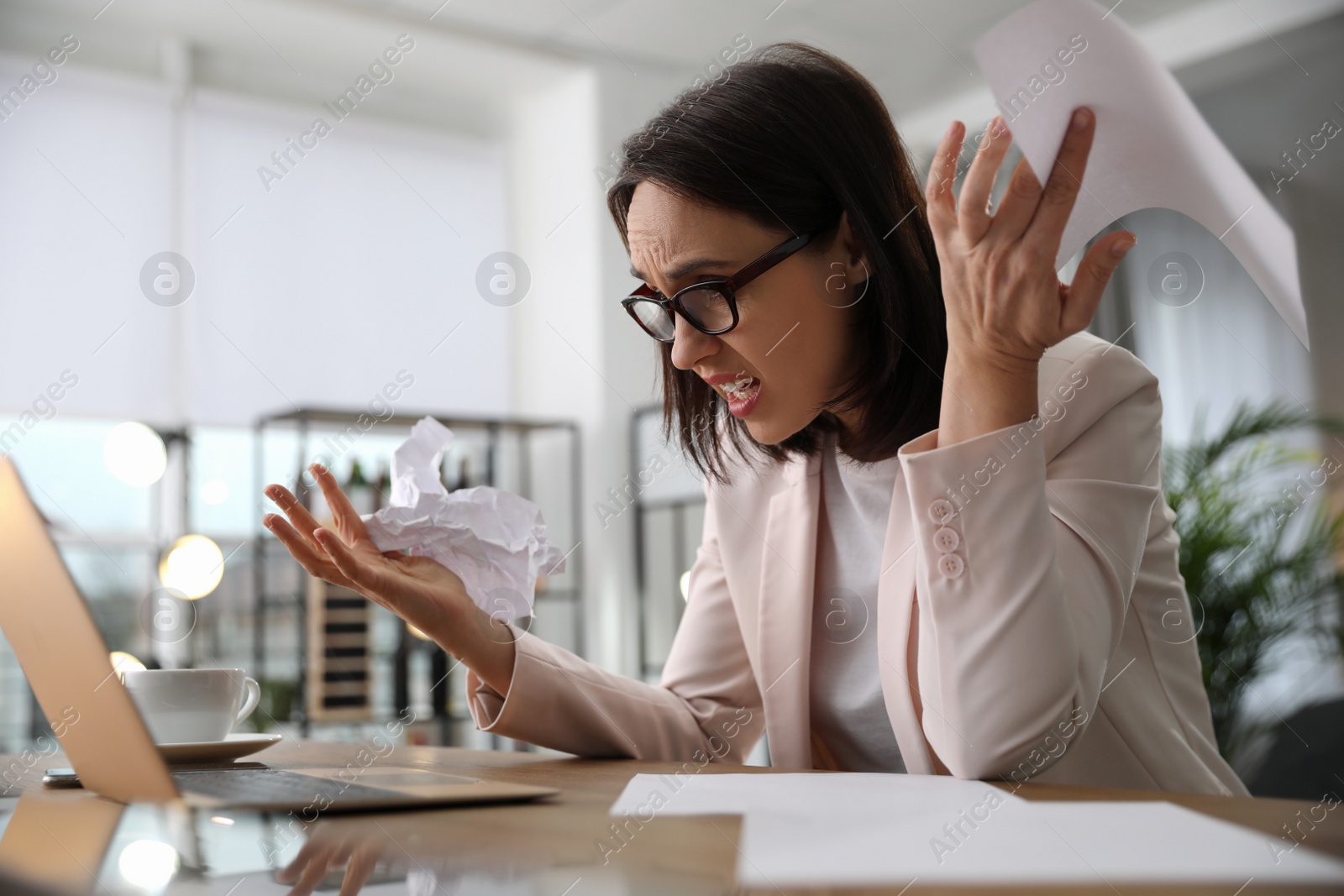 Photo of Emotional businesswoman with crumpled paper working on laptop in office. Online hate concept