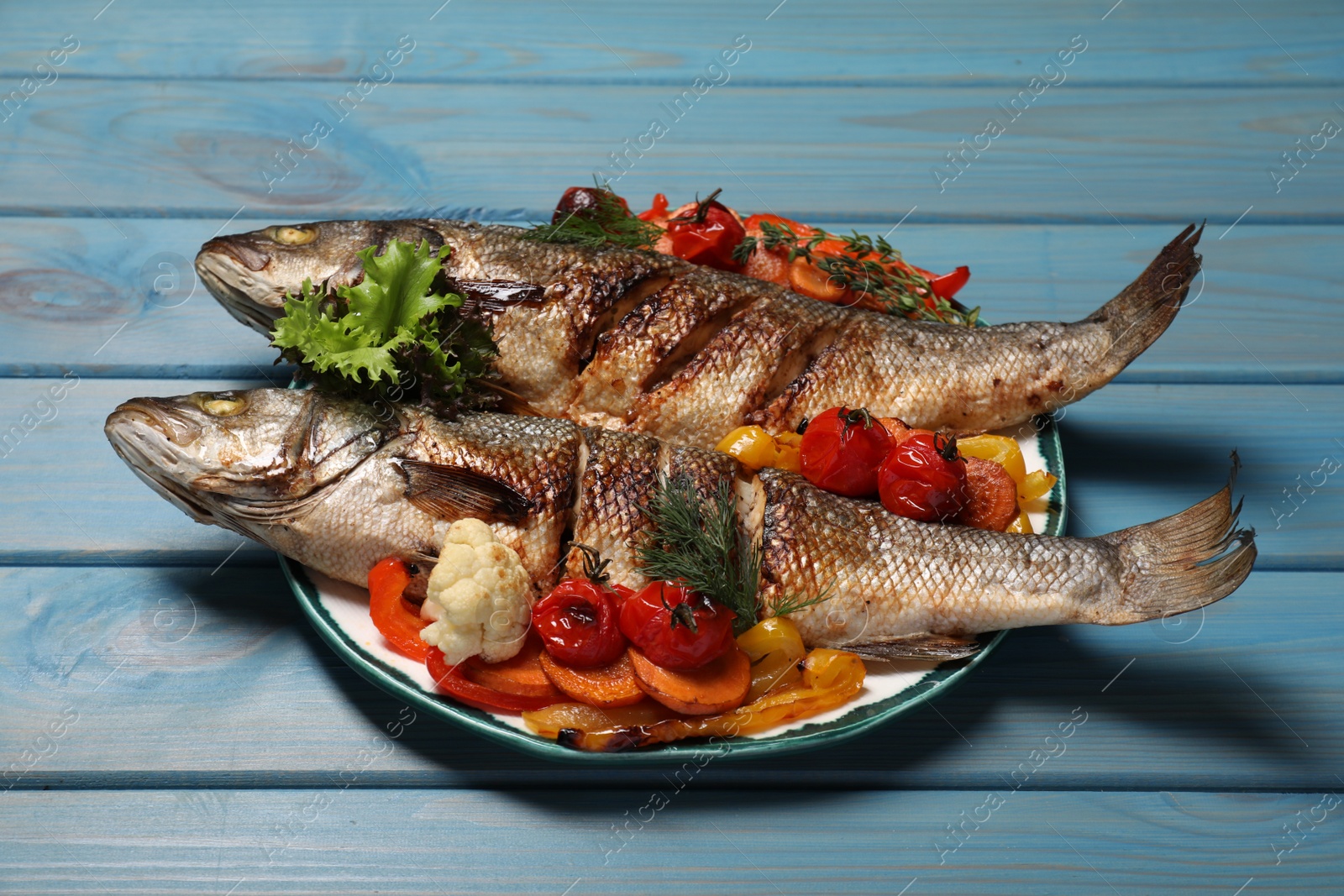 Photo of Plate with delicious baked sea bass fish and vegetables on light blue table