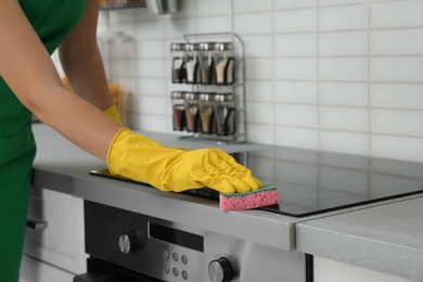 Photo of Female janitor cleaning kitchen stove with sponge, closeup
