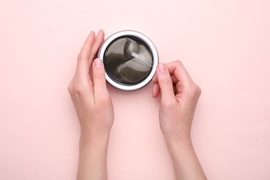 Photo of Woman taking under eye patch with spatula out of jar on light pink background, top view. Cosmetic product