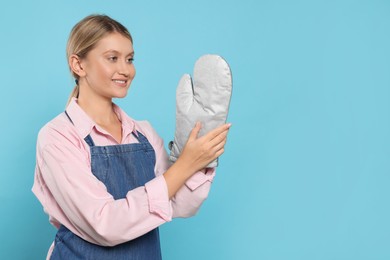 Photo of Beautiful young woman in denim apron and oven glove on light blue background, closeup. Space for text