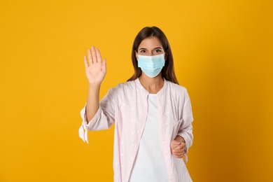 Photo of Woman in protective mask showing hello gesture on yellow background. Keeping social distance during coronavirus pandemic