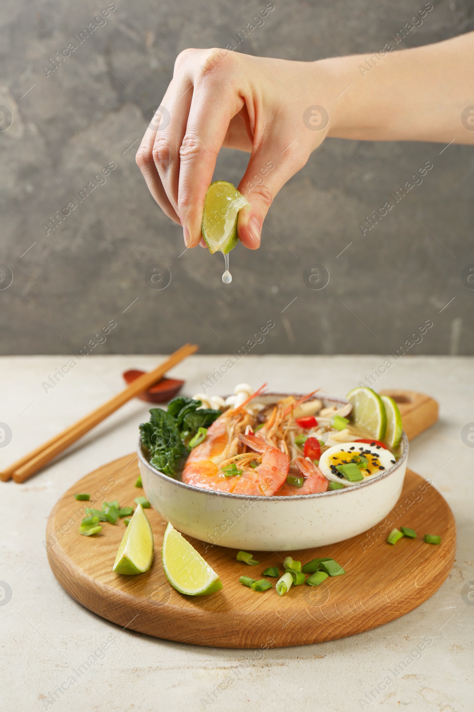 Photo of Woman squeezing lime juice into bowl of delicious ramen with shrimps at light textured table, closeup. Noodle soup