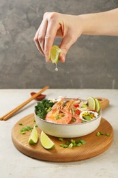 Woman squeezing lime juice into bowl of delicious ramen with shrimps at light textured table, closeup. Noodle soup