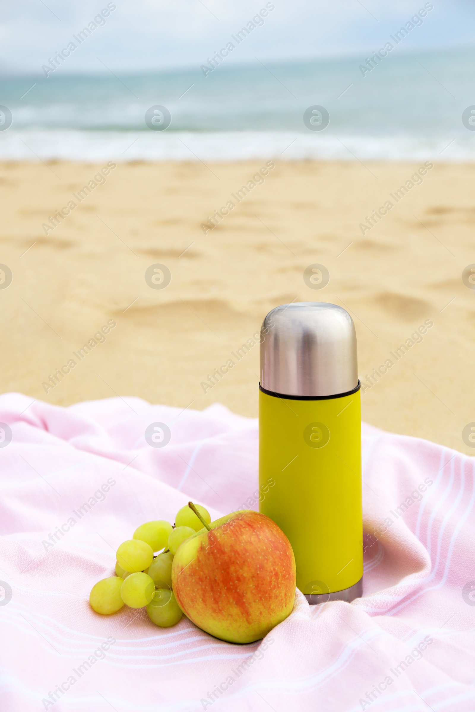 Photo of Metallic thermos with hot drink, fruits and plaid on sandy beach near sea