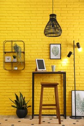 Photo of High wooden table with tablet and stool as stand up workplace near yellow brick wall. Stylish interior