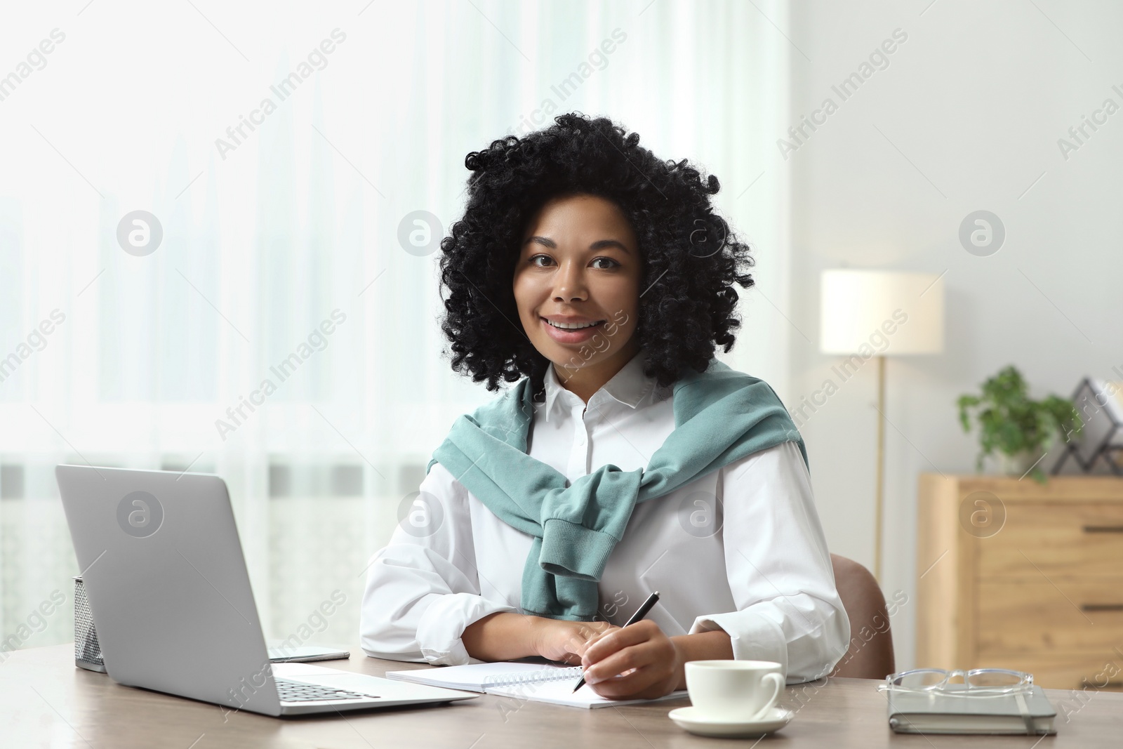 Photo of Happy young woman writing notes while using laptop at wooden desk indoors