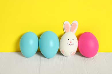 Photo of Bright Easter eggs and white one as cute bunny on wooden table against yellow background
