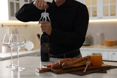 Romantic dinner. Man opening wine bottle with corkscrew at table in kitchen, closeup