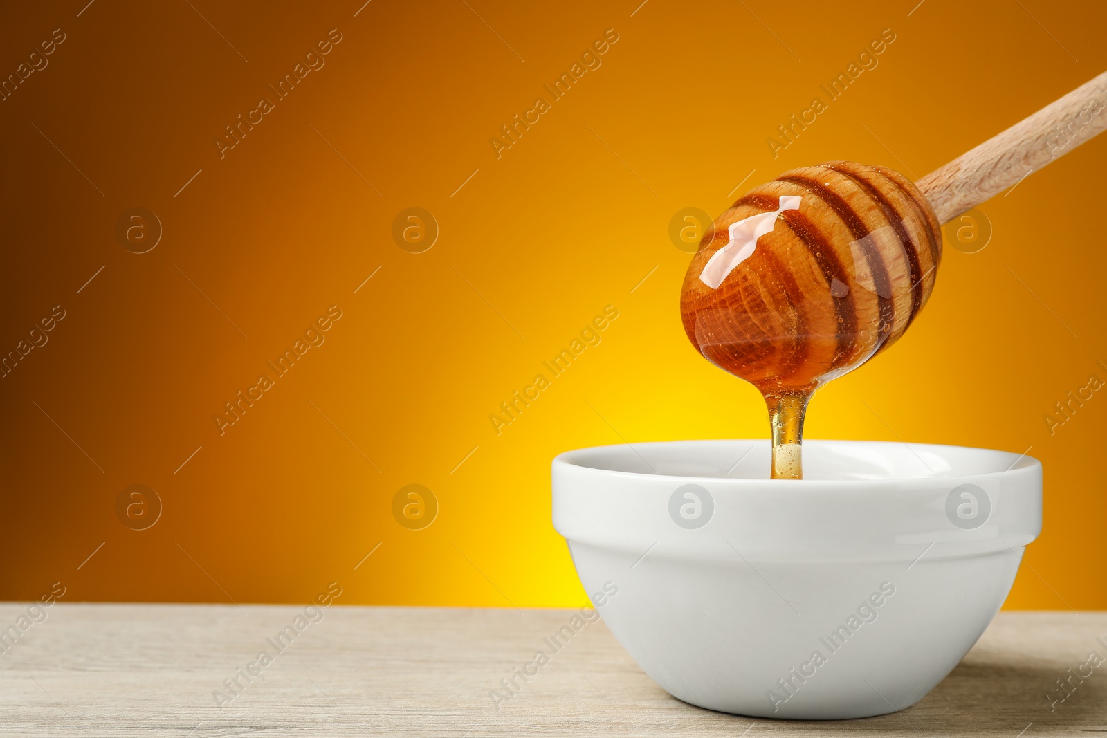 Photo of Pouring honey from dipper into bowl at wooden table against golden background, space for text