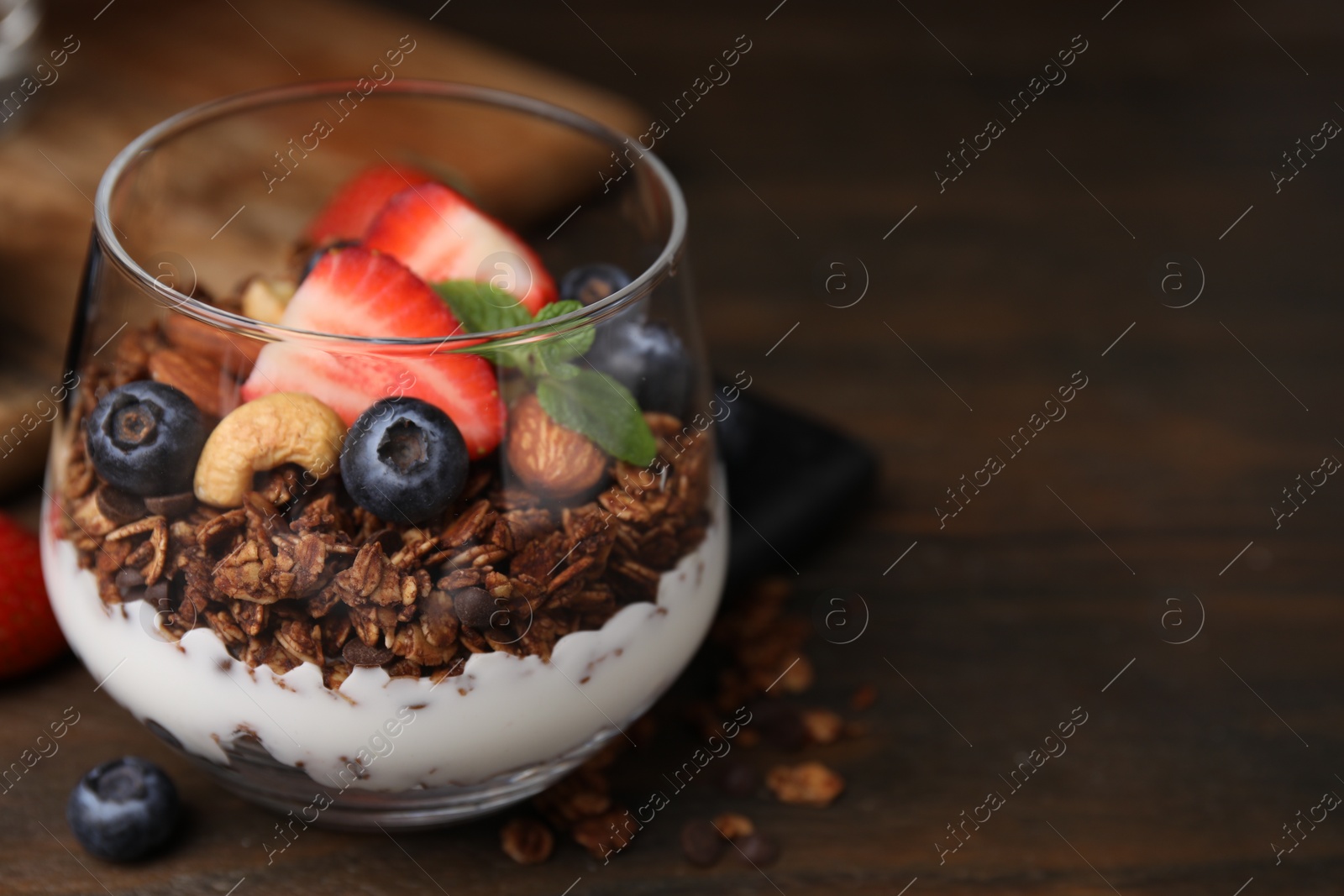 Photo of Tasty granola with berries, nuts and yogurt in glass on wooden table, closeup. Space for text