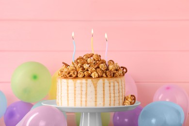 Photo of Caramel drip cake decorated with popcorn and pretzels near balloons against pink wooden background