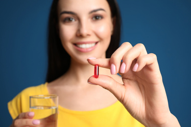 Young woman with glass of water and vitamin capsule against blue background, focus on hand